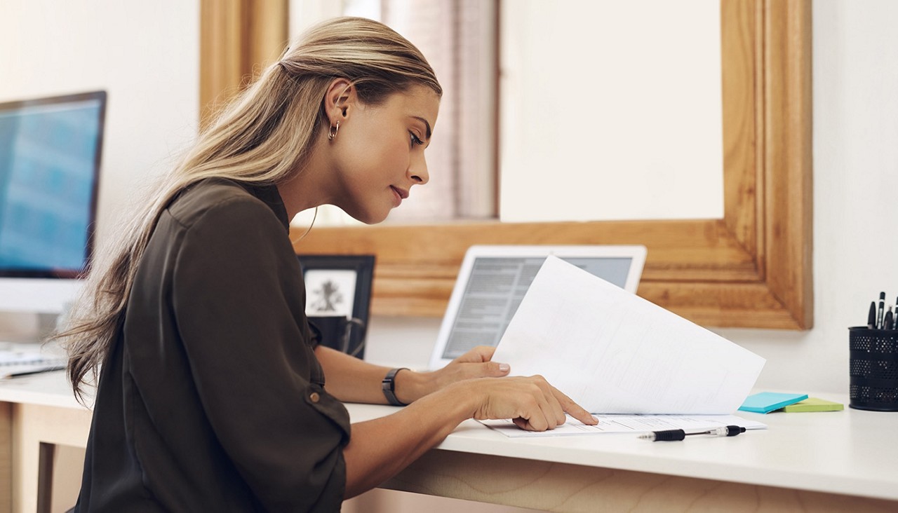 A woman in front of a laptop looking over paperwork.