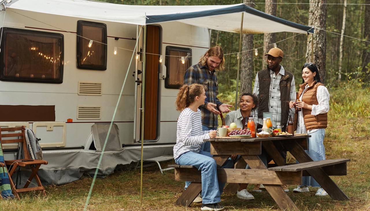 A group of young people enjoy a picnic outdoors next to their camping trailer