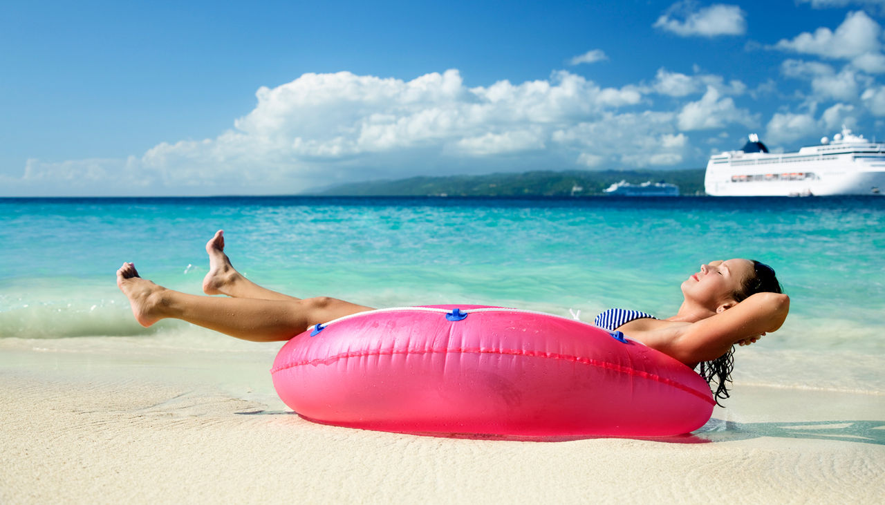 Woman sunbathing on a pink inner tube on a beach with a cruise ship in the background
